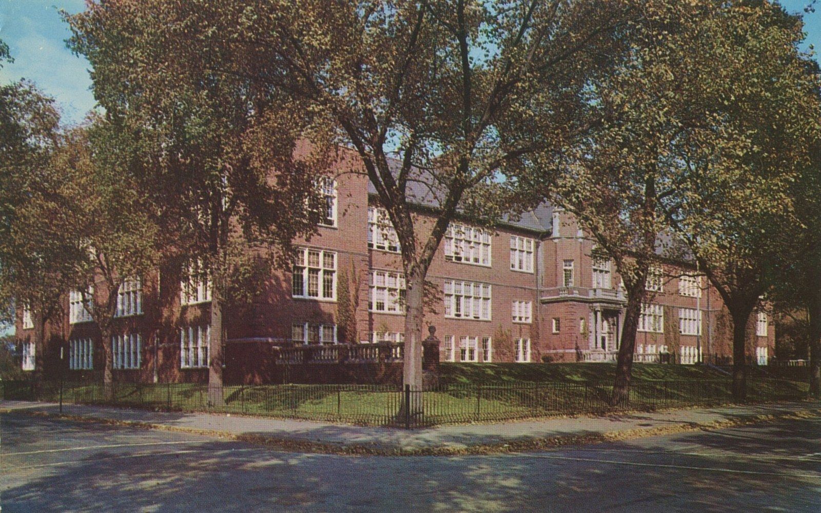 Brick school building with arched windows, fronted by trees and a low iron fence