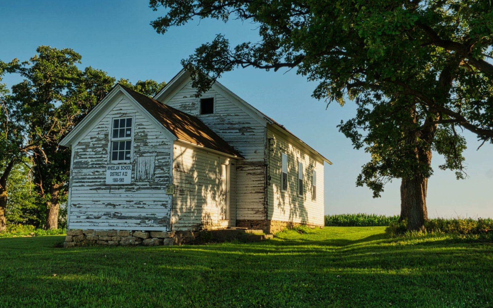 Weathered one-room schoolhouse with peeling white paint stands beneath a shady oak tree
